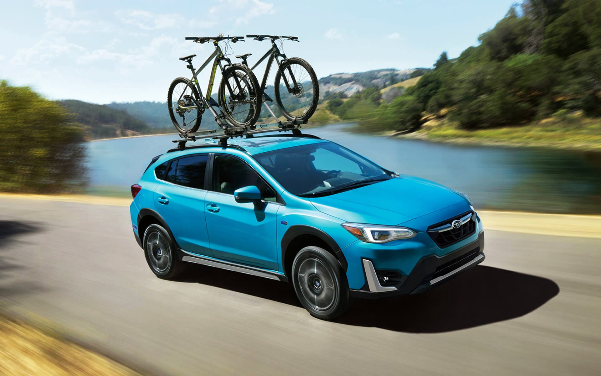 A blue Crosstrek Hybrid with two bicycles on its roof rack driving beside a river | Sierra Subaru of Monrovia in Monrovia CA